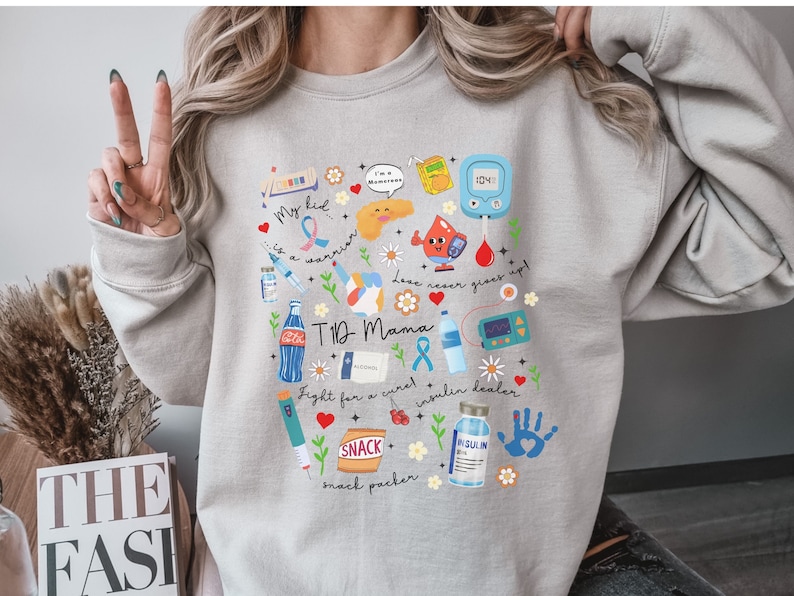 Op de afbeelding: Een lichtgrijs sweatshirt met een kleurrijk grafisch ontwerp met een pancreas, een insulinepomp, een bloedglucosemeter en andere diabetesgerelateerde afbeeldingen. De tekst op het sweatshirt luidt "My kid is a warrior", "T1D Mama", "Love never gives up", "Fight for a cure", "Insulin dealer" en "Snack packer".