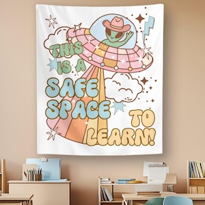 May include: A white wall tapestry featuring a green alien wearing a pink cowboy hat inside a flying saucer. The design includes clouds, stars, and a lightning bolt with the text THIS IS A SAFE SPACE TO LEARN in blue, pink, and yellow bubble letters.