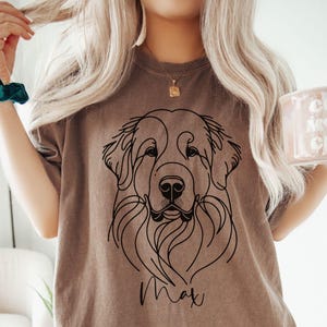 Op de afbeelding: Een bruin T-shirt met een zwarte lijntekening van een hondenkop en de naam "Max" gedrukt op de voorkant.