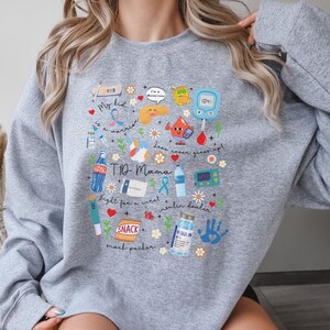 Op de afbeelding: Grijs sweatshirt met een kleurrijk grafisch ontwerp met verschillende diabetes-gerelateerde pictogrammen, waaronder een bloedglucosemeter, insulineflacons en een snackpakket. De tekst "T1D Mama" staat op de grafiek.