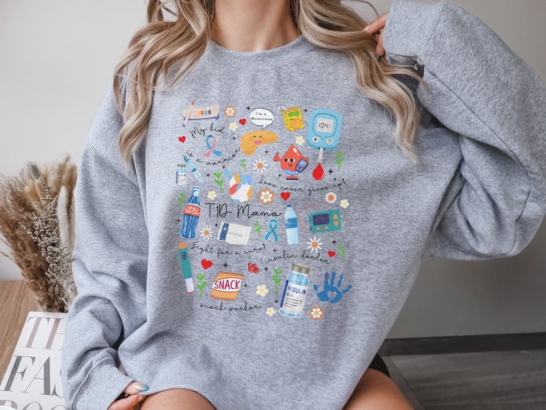 Op de afbeelding: Grijs sweatshirt met een kleurrijk grafisch ontwerp met verschillende diabetes-gerelateerde pictogrammen, waaronder een bloedglucosemeter, insulineflacons en een snackpakket. De tekst "T1D Mama" staat op de grafiek.