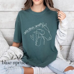 Op de afbeelding: Een tealblauw t-shirt met een witte lijntekening van een vrouw met een cowboyhoed die een paard berijdt. De tekst "Hold Your Horses" staat boven de tekening.