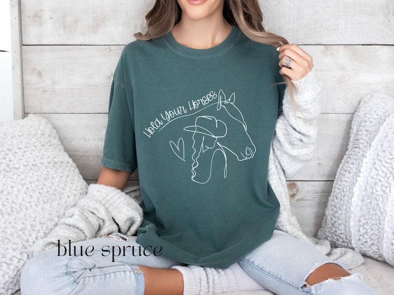 Op de afbeelding: Een tealblauw t-shirt met een witte lijntekening van een vrouw met een cowboyhoed die een paard berijdt. De tekst "Hold Your Horses" staat boven de tekening.