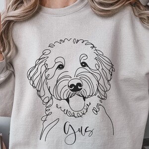 May include: A light gray sweatshirt featuring a black line drawing of a dog's face, with the name "Gus" in cursive below. The dog's features are detailed with continuous lines. The sweatshirt is a neutral color.