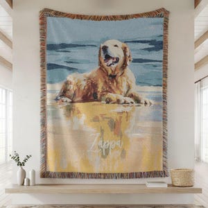 May include: Woven throw blanket featuring a golden retriever dog lying on a beach. The dog is depicted in shades of gold, brown, and white, with a blue and yellow background. The name "Zappa" is written on the blanket.
