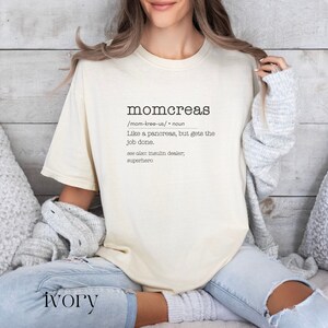 May include: A beige t-shirt with the word "momcreas" defined as "Like a pancreas, but gets the job done." The text is in black and the shirt is worn by a person sitting on a couch.