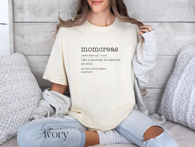 May include: A beige t-shirt with the word "momcreas" defined as "Like a pancreas, but gets the job done." The text is in black and the shirt is worn by a person sitting on a couch.