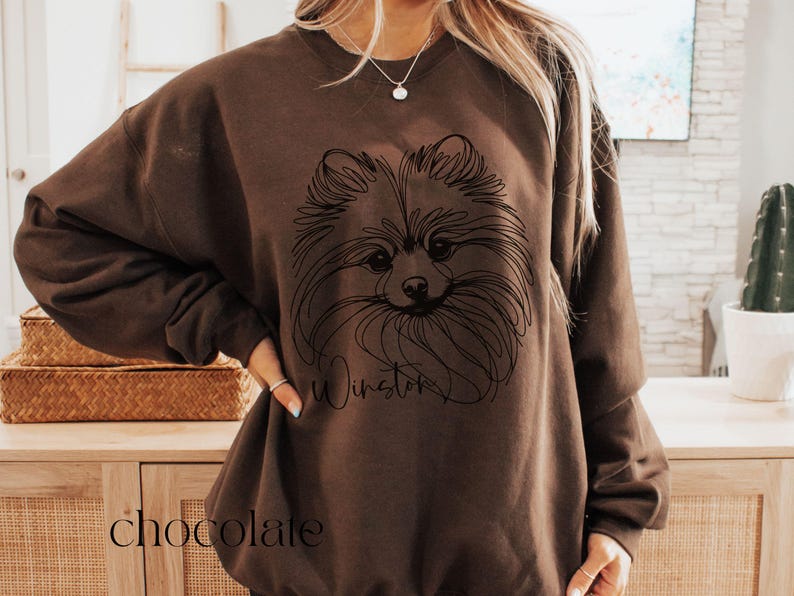 May include: Chocolate-colored sweatshirt featuring a line drawing of a Pomeranian dog's face. The dog's name, "Winston," is written below the image in a script font. The word "chocolate" is printed at the bottom of the sweatshirt.