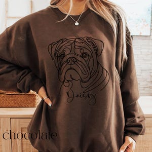 K&ouml;nnte beinhalten: Schokoladenfarbenes Sweatshirt mit einer Strichzeichnung eines Bulldoggen-Gesichts. Das Wort "Daisy" ist in Kursivschrift unter dem Bild des Hundes geschrieben. Das Wort "chocolate" ist unten auf dem Sweatshirt aufgedruckt.