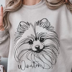 May include: A light gray sweatshirt featuring a black line drawing of a Pomeranian dog's face. The dog's name, "Winston," is written in cursive below the image. The sweatshirt is a neutral color.