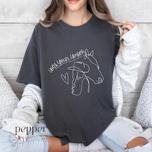 Op de afbeelding: Een donkergrijs T-shirt met een witte lijntekening van een vrouw die een cowboyhoed draagt en een paardenkop vasthoudt. De tekst "Hold Your Horses" staat boven de tekening.