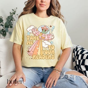 Puede incluir: Camiseta amarilla mantequilla con un gr&aacute;fico retro de un extraterrestre con sombrero de vaquero dentro de un OVNI, irradiando un arco&iacute;ris. El texto dice "Take Me To Your Readers". La camiseta es de corte holgado.