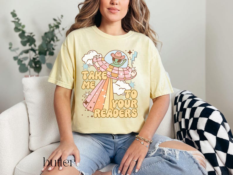 Puede incluir: Camiseta amarilla mantequilla con un gr&aacute;fico retro de un extraterrestre con sombrero de vaquero dentro de un OVNI, irradiando un arco&iacute;ris. El texto dice "Take Me To Your Readers". La camiseta es de corte holgado.