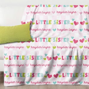 May include: A white blanket with the words "LITTLE SISTER" in colorful letters, interspersed with hearts and the name "hayden layne". A lime green pillow rests on a white sofa.