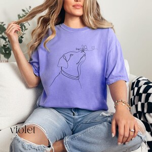 Op de afbeelding: Violetkleurig T-shirt met een minimalistische lijntekening van een hond die naar een vlinder kijkt. Het shirt heeft korte mouwen en een relaxte pasvorm. Het woord "keeps" staat boven de kop van de hond en een hartje is naast de vlinder getekend.