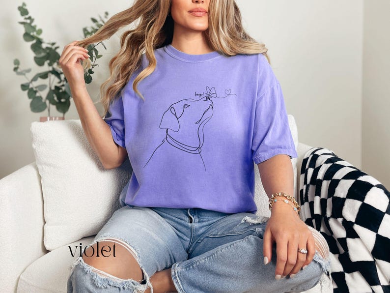 Op de afbeelding: Violetkleurig T-shirt met een minimalistische lijntekening van een hond die naar een vlinder kijkt. Het shirt heeft korte mouwen en een relaxte pasvorm. Het woord "keeps" staat boven de kop van de hond en een hartje is naast de vlinder getekend.