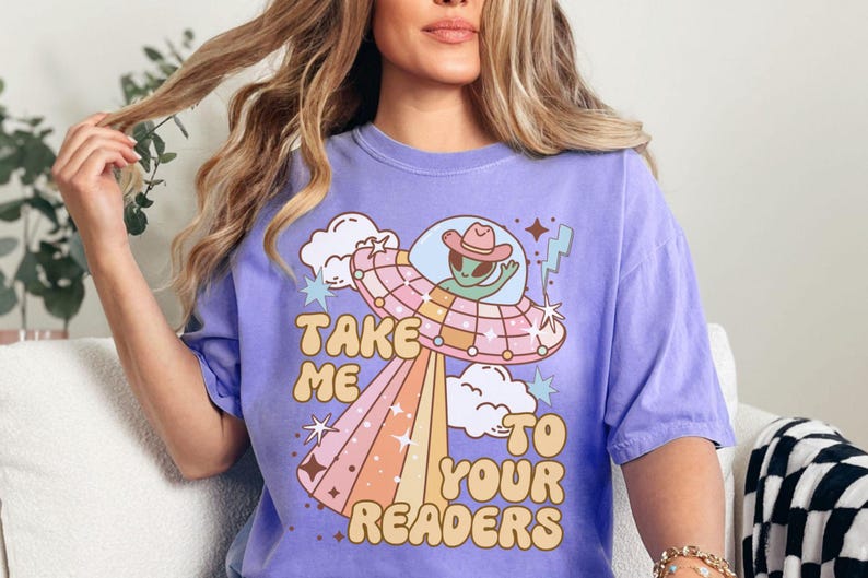 Puede incluir: Camiseta lila con un dise&ntilde;o retro de un extraterrestre con sombrero de vaquero dentro de un platillo volante. El texto "Take Me To Your Readers" est&aacute; impreso en una fuente juguetona, con rayos de arco&iacute;ris pastel que se extienden desde el platillo.