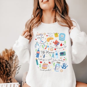 Op de afbeelding: Wit sweatshirt met een kleurrijk grafisch ontwerp met verschillende symbolen die te maken hebben met diabetes, waaronder een bloedglucosemeter, een insulinepomp en een spuit. De tekst "T1D Mama" is prominent op het sweatshirt afgebeeld.