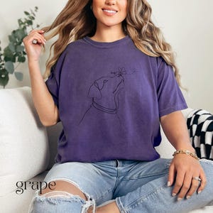 Op de afbeelding: Een paars T-shirt met een lijntekening van een hond die naar een vlinder kijkt. Het woord "hope" staat boven de kop van de hond. Het woord "grape" staat linksonder in de afbeelding.