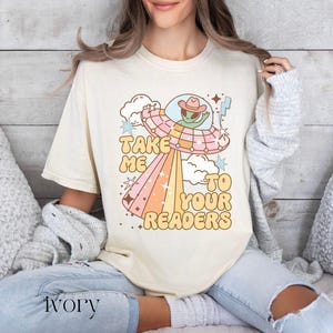 Puede incluir: Camiseta color marfil con un dise&ntilde;o retro de un OVNI y un extraterrestre con sombrero de vaquero. El texto "TAKE ME TO YOUR READERS" est&aacute; en una fuente divertida. La persona lleva vaqueros claros. La palabra "ivory" est&aacute; impresa abajo.