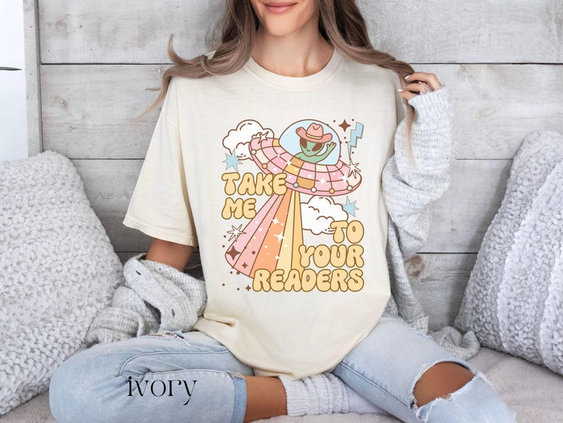 Puede incluir: Camiseta color marfil con un dise&ntilde;o retro de un OVNI y un extraterrestre con sombrero de vaquero. El texto "TAKE ME TO YOUR READERS" est&aacute; en una fuente divertida. La persona lleva vaqueros claros. La palabra "ivory" est&aacute; impresa abajo.