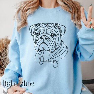 K&ouml;nnte beinhalten: Hellblaues Sweatshirt mit einer schwarzen Strichzeichnung eines Bulldoggen-Gesichts und dem Namen "Daisy" in Schreibschrift. Der Text "light blue" ist unten aufgedruckt. Das Sweatshirt hat lange &Auml;rmel.