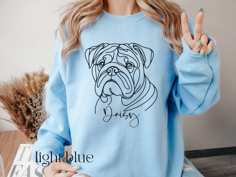K&ouml;nnte beinhalten: Hellblaues Sweatshirt mit einer schwarzen Strichzeichnung eines Bulldoggen-Gesichts und dem Namen "Daisy" in Schreibschrift. Der Text "light blue" ist unten aufgedruckt. Das Sweatshirt hat lange &Auml;rmel.