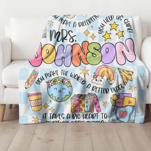 May include: A blue and white checkered fleece blanket with a colorful design featuring the text "You make a difference", "Mrs. Johnson", "You help us shine", "You make the world a better place", and "It takes a big heart to do what we do".