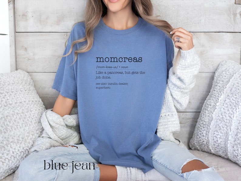 May include: A blue t-shirt with the word "momcreas" defined as "like a pancreas, but gets the job done." The text is in black and the shirt is worn by a person sitting on a couch.