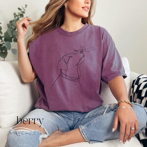 Op de afbeelding: Bessenkleurig T-shirt met een lijntekening van een hond die naar een vlinder kijkt. Het woord "Berry" staat onderaan gedrukt. Het shirt heeft korte mouwen en een relaxte pasvorm.