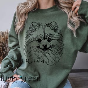 May include: Military green sweatshirt featuring a black line drawing of a Pomeranian dog's face. The dog's name, "Winston," is written in cursive below the drawing. The words "Military green" are printed at the bottom of the sweatshirt.