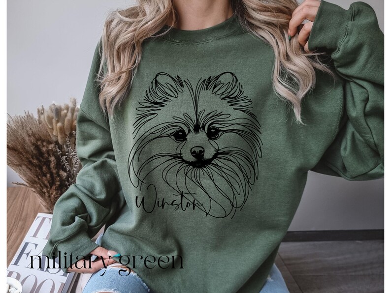 May include: Military green sweatshirt featuring a black line drawing of a Pomeranian dog's face. The dog's name, "Winston," is written in cursive below the drawing. The words "Military green" are printed at the bottom of the sweatshirt.