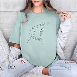 Op de afbeelding: Lichtgroen T-shirt met een minimalistische lijntekening van een hond die naar een vlinder kijkt. Het woord "bay" is in scriptletter onderaan links gedrukt. Het shirt heeft een losse pasvorm.
