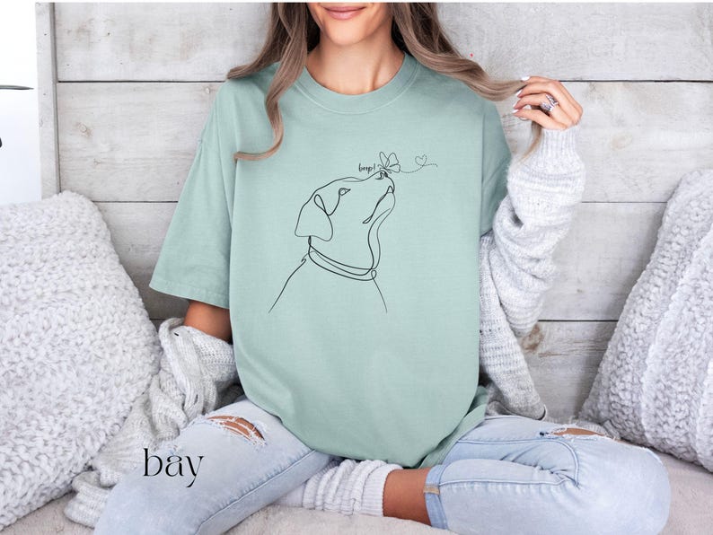 Op de afbeelding: Lichtgroen T-shirt met een minimalistische lijntekening van een hond die naar een vlinder kijkt. Het woord "bay" is in scriptletter onderaan links gedrukt. Het shirt heeft een losse pasvorm.