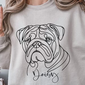 K&ouml;nnte beinhalten: Ein hellgraues Sweatshirt mit einer schwarzen Strichzeichnung des Gesichts einer Bulldogge. Der Name des Hundes, "Daisy", ist unter dem Bild in Schreibschrift geschrieben. Die Person auf dem Bild tr&auml;gt das Sweatshirt und h&auml;lt zwei Finger hoch.