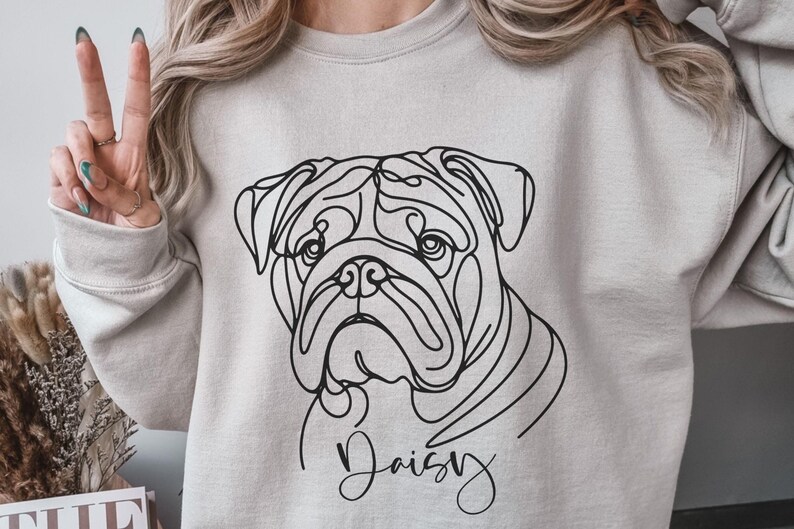 K&ouml;nnte beinhalten: Ein hellgraues Sweatshirt mit einer schwarzen Strichzeichnung des Gesichts einer Bulldogge. Der Name des Hundes, "Daisy", ist unter dem Bild in Schreibschrift geschrieben. Die Person auf dem Bild tr&auml;gt das Sweatshirt und h&auml;lt zwei Finger hoch.