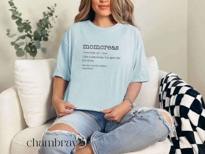 May include: A light blue t-shirt with the word "momcreas" defined as "Like a pancreas, but gets the job done." The text is in black and the shirt is on a person sitting on a white couch.