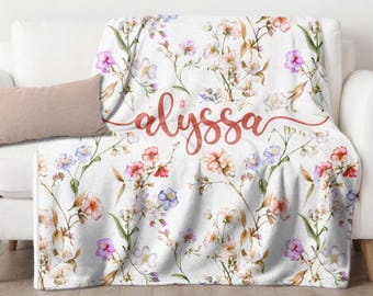 Puede incluir: Una manta floral blanca con un diseño de acuarela de flores rosas, moradas y azules. La manta está sobre un sofá blanco con un cojín a juego. El nombre "alyssa" está escrito en cursiva sobre la manta.