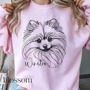 May include: A light pink sweatshirt featuring a black line drawing of a Pomeranian dog's face. The dog's name, "Winston," is written in cursive below the image. The word "blossom" is visible in the lower left corner.