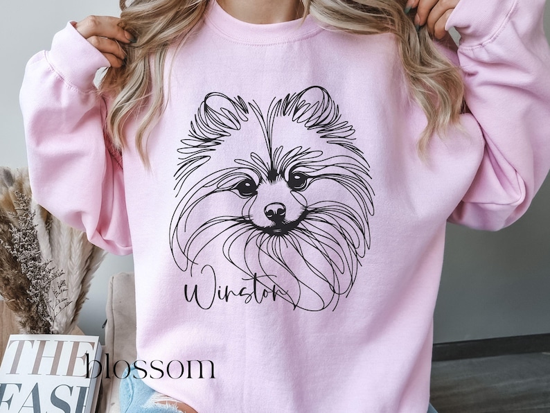 May include: A light pink sweatshirt featuring a black line drawing of a Pomeranian dog's face. The dog's name, "Winston," is written in cursive below the image. The word "blossom" is visible in the lower left corner.
