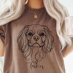 May include: A brown t-shirt featuring a black line drawing of a Cavalier King Charles Spaniel dog. The dog's name, "Tucker," is written in cursive below the image. A pink mug with white text is held in the right hand.