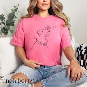 Op de afbeelding: Een roze t-shirt met een lijntekening van een hond die naar een vlinder en twee harten kijkt. Het woord "boop!" staat boven de hond. De tekst "crunch berry" staat onderaan de afbeelding.