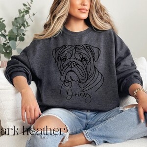 K&ouml;nnte beinhalten: Dunkelgraues Sweatshirt mit einer Strichzeichnung eines Bulldoggen-Gesichts und dem Namen "Daisy" darunter. Der Text "dark heather" ist unten aufgedruckt. Ein l&auml;ssiger, bequemer Stil.