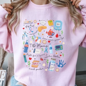 Op de afbeelding: Roze trui met een grafisch ontwerp met verschillende illustraties met betrekking tot type 1 diabetes. Het ontwerp bevat een pancreas, een insulinepomp, een bloedglucosemeter en andere medische benodigdheden. De tekst op de trui luidt "My kid is a warrior", "T1D Mama", "Fight for a cure", "Insulin dealer", "Snack packer" en "Love never gives up".