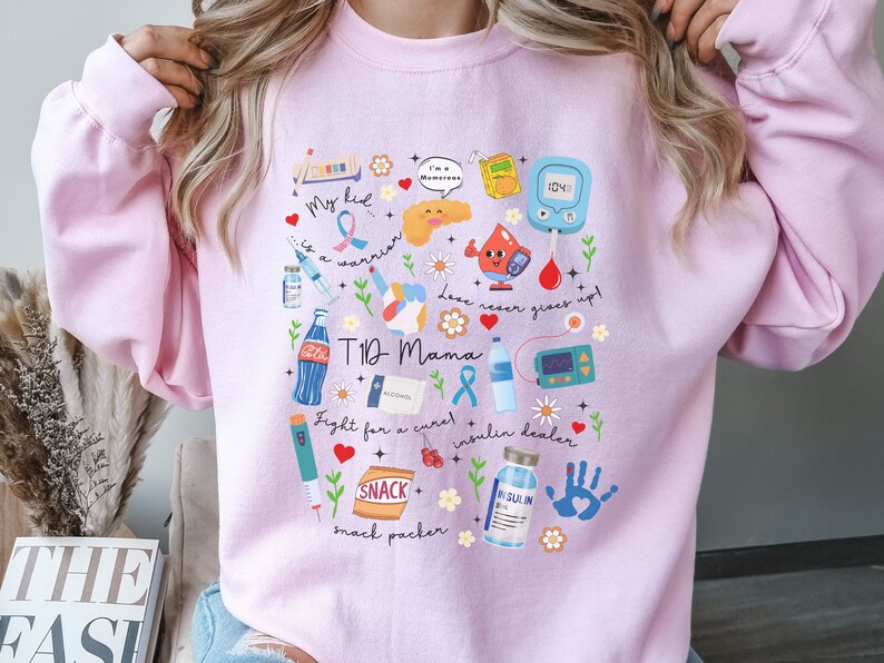 Op de afbeelding: Roze trui met een grafisch ontwerp met verschillende illustraties met betrekking tot type 1 diabetes. Het ontwerp bevat een pancreas, een insulinepomp, een bloedglucosemeter en andere medische benodigdheden. De tekst op de trui luidt "My kid is a warrior", "T1D Mama", "Fight for a cure", "Insulin dealer", "Snack packer" en "Love never gives up".