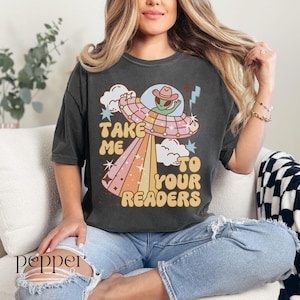 Puede incluir: Camiseta gris oscuro con un gr&aacute;fico retro de un extraterrestre con sombrero de vaquero dentro de un OVNI, con el texto "Take Me To Your Readers" en una fuente vintage. El dise&ntilde;o presenta colores pastel y estrellas.