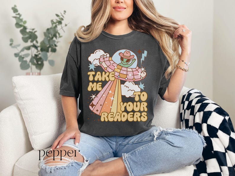 Puede incluir: Camiseta gris oscuro con un gr&aacute;fico retro de un extraterrestre con sombrero de vaquero dentro de un OVNI, con el texto "Take Me To Your Readers" en una fuente vintage. El dise&ntilde;o presenta colores pastel y estrellas.