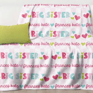 May include: A white blanket with a pink and white checkered pattern, featuring the words "BIG SISTER" in teal and pink, along with hearts and the name "frances kate". A lime green pillow rests on a white sofa.