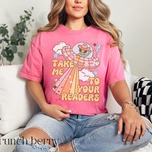 Puede incluir: Camiseta rosa con el texto "Take Me To Your Readers" y un gr&aacute;fico de un extraterrestre con sombrero de vaquero en un OVNI. El dise&ntilde;o incluye un arco&iacute;ris y nubes. La camiseta se lleva con vaqueros azules.