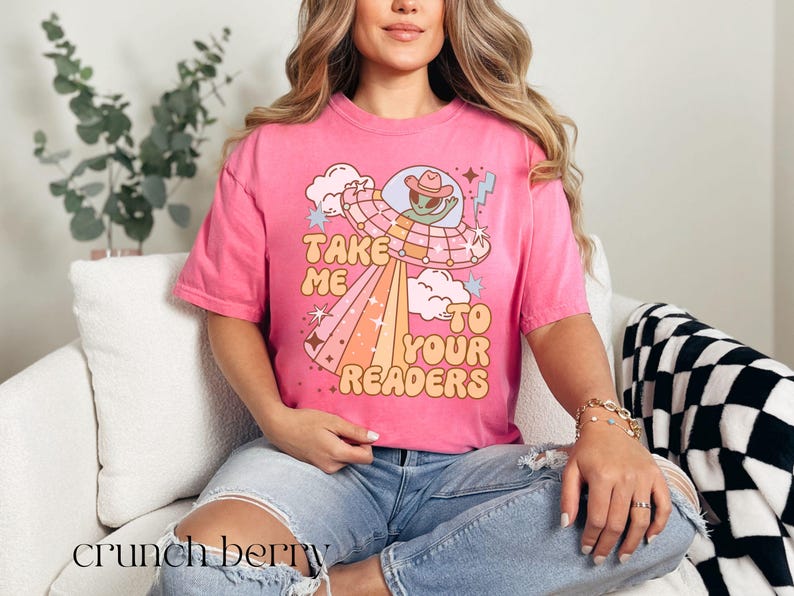 Puede incluir: Camiseta rosa con el texto "Take Me To Your Readers" y un gr&aacute;fico de un extraterrestre con sombrero de vaquero en un OVNI. El dise&ntilde;o incluye un arco&iacute;ris y nubes. La camiseta se lleva con vaqueros azules.